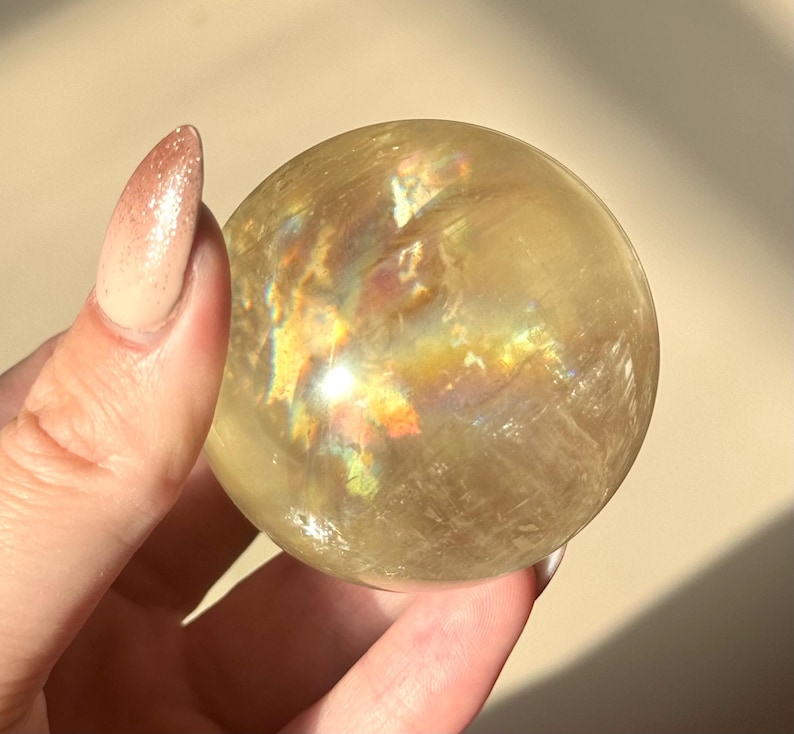 Op de afbeelding: Een doorschijnende, goudgele kristallen bol met iriserende regenboogreflecties. Het gepolijste oppervlak van de bol reflecteert het licht. De kristallen bol wordt in een hand gehouden. Een decoratief item.