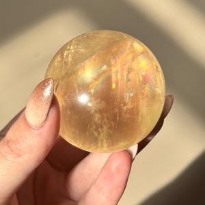Op de afbeelding: Een heldere, goudgele kristallen bol, vastgehouden in een hand. De bol heeft interne regenboogachtige brekingen. De achtergrond is een neutrale beige kleur, met schaduwen. De bol is een decoratief item.