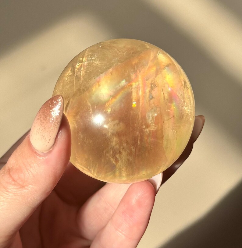 Op de afbeelding: Een heldere, goudgele kristallen bol, vastgehouden in een hand. De bol heeft interne regenboogachtige brekingen. De achtergrond is een neutrale beige kleur, met schaduwen. De bol is een decoratief item.