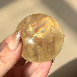 Op de afbeelding: Een heldere, goudgele kristallen bol. De bol heeft interne breuken en een regenboogglans. De kristallen bol wordt vastgehouden in een hand met een lichtgekleurde manicure. De achtergrond is een zachte, neutrale tint.