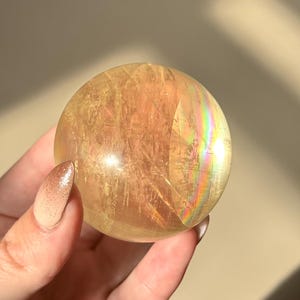 Op de afbeelding: Een heldere, goudgele kristallen bol met interne breuken en regenboogirisatie. De bol wordt in een hand gehouden, waarbij het gepolijste oppervlak wordt getoond. Een decoratief en uniek item.