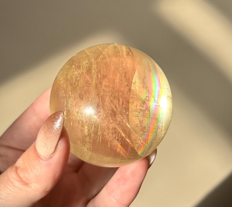 Op de afbeelding: Een heldere, goudgele kristallen bol met interne breuken en regenboogirisatie. De bol wordt in een hand gehouden, waarbij het gepolijste oppervlak wordt getoond. Een decoratief en uniek item.