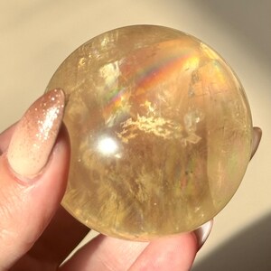 Op de afbeelding: Een doorschijnende, goudgele kristallen bol. De bol heeft interne patronen en een regenboogachtige iriserende glans. De kristal wordt vastgehouden in een hand met een lichtgekleurde manicure.
