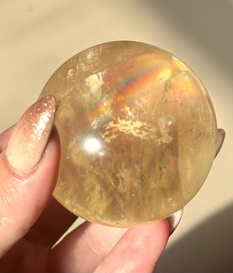 Op de afbeelding: Een doorschijnende, goudgele kristallen bol. De bol heeft interne patronen en een regenboogachtige iriserende glans. De kristal wordt vastgehouden in een hand met een lichtgekleurde manicure.