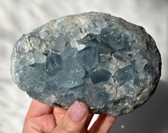 May include: A large, rough-hewn celestite crystal cluster. The crystal features a mix of light blue and gray hues, with a crystalline structure. The stone is held in a hand, showcasing its size and texture. The overall aesthetic is natural and earthy.