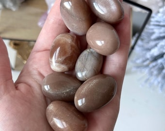 May include: A collection of seven sunstone tumbled stones in various shades of brown and beige. The stones are smooth and polished, with a slight shimmer.