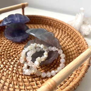 May include: Two amethyst crystal clam shell boxes with a white moonstone bracelet inside one of the shells. The shells are sitting on a woven wicker tray with a wooden handle.