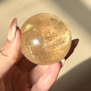 Op de afbeelding: Een goudgele, doorschijnende kristallen bol. De bol wordt in een hand gehouden, waardoor het gladde, gepolijste oppervlak zichtbaar wordt. Het kristal heeft interne strepen en een zachte, natuurlijke gloed.