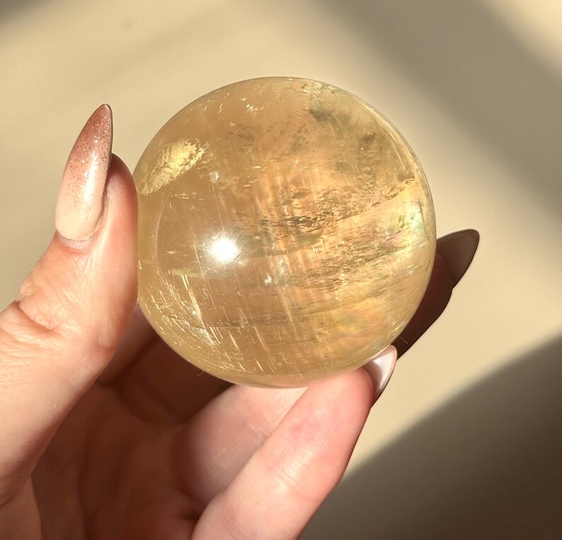 Op de afbeelding: Een goudgele, doorschijnende kristallen bol. De bol wordt in een hand gehouden, waardoor het gladde, gepolijste oppervlak zichtbaar wordt. Het kristal heeft interne strepen en een zachte, natuurlijke gloed.