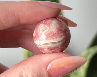 Könnte beinhalten: Eine rosa und weiße Kugel aus poliertem rosa Calcit, eine Kristallsorte, die für ihre beruhigende Energie bekannt ist.