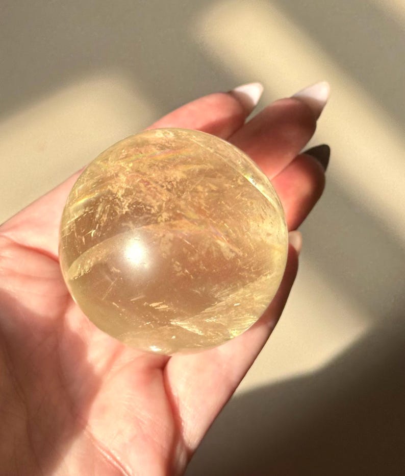 Op de afbeelding: Een heldere, goudgele kristallen bol. De bol wordt in een hand gehouden, met licht dat weerkaatst op het oppervlak. Het kristal heeft interne patronen en insluitsels, wat het een unieke uitstraling geeft. De algehele esthetiek is natuurlijk en aards.