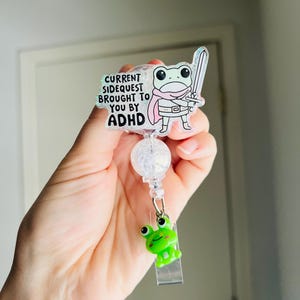 May include: A clear acrylic badge reel with a cartoon frog holding a sword. The badge reel has the text "CURRENT SIDEQUEST BROUGHT TO YOU BY ADHD". A small green frog charm is attached to the reel.
