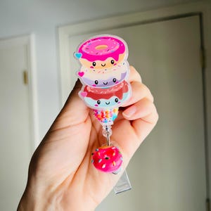 May include: A stack of cartoon donuts in pink, purple, and red with smiling faces and heart accents. The badge reel has a pink donut charm at the end. The donuts are on a clear badge reel.