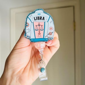 May include: A badge reel featuring a Libra zodiac design. The badge has a light blue jacket shape with the word "LIBRA" at the top. The design includes a scale symbol, stars, and a rose. The reel has a glittery pink top and a clear plastic clip.