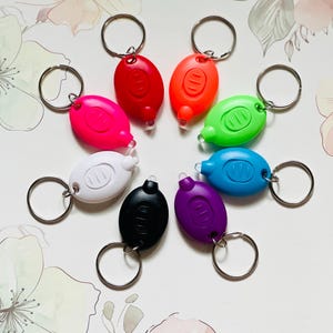 May include: A collection of eight small, oval-shaped LED keychains in various colors: pink, red, orange, green, white, black, purple, and blue. Each keychain has a silver ring and a small button.