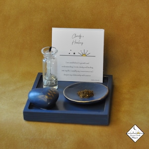 May include: A blue wooden tray with a small glass bottle, a blue heart-shaped stone, a small blue dish with brown powder, and a white card with the text "Clarity & Healing" and a sun and stars graphic. The card reads "I am established in growth and understanding. I invite clarity and healing into my life. I amplify my resonance and deepen my relationship with source."