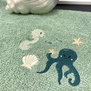 Könnte beinhalten: Ein grünes Handtuch mit gestickten Meeresbewohnern, darunter ein Oktopus, ein Seepferdchen und eine Muschel. Im Hintergrund befindet sich eine weiße Muschel mit irisierenden Farben.