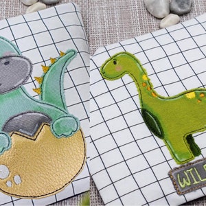 Può includere: Due disegni di dinosauri ricamati su tessuto bianco con un motivo a griglia. Il primo disegno è un dinosauro verde che esce da un uovo dorato. Il secondo disegno è un dinosauro verde con il testo "Wildfang" sotto.