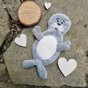 May include: A gray plush seal toy with a white belly and embroidered facial features. The seal is lying on a gray stone surface with wooden hearts scattered around it. The text "Plotta Kreativ" is engraved on a wooden round.