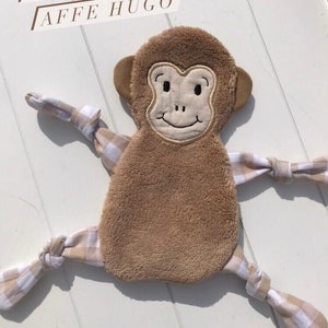 Puede incluir: Un juguete de mono de peluche marrón con una tela a cuadros blancos y marrones atada a sus brazos y piernas. El mono tiene una cara sonriente con ojos y nariz bordados. El texto "IN THE HOOP" y "AFFE HUGO" está impreso sobre el mono.