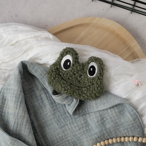 May include: A green, plush frog face with white eyes and black pupils rests on a gray, hooded blanket. The frog has a smiling expression. The blanket is draped over a wooden tray and white fabric.