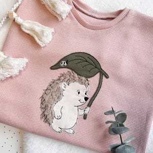 Peut inclure: Un t-shirt à manches longues rose clair en maille gaufrée avec un hérisson brodé tenant une feuille. Le hérisson a un corps blanc et un dos marron texturé. La feuille est vert olive. Le t-shirt a une encolure ronde.