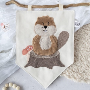 May include: A cream-colored banner with a brown beaver applique. The beaver is sitting on a tree stump with a mushroom. The banner is suspended by a wooden stick and cream-colored fabric loops.