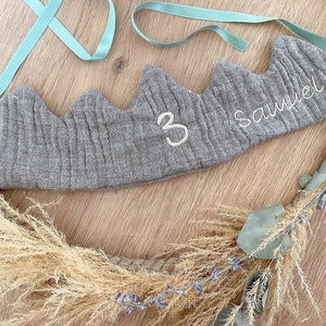 Könnte beinhalten: Ein graues Stoffkrönchen mit bergähnlichem Design, bestickt mit dem Namen "Samuel" und der Zahl "3" in Weiß. Die Krone hat hellblaue Bänder und ist Teil eines dekorativen Kranzes mit Trockenblumen und Seil.