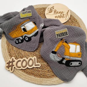 May include: Two grey muslin triangle bibs featuring embroidered construction vehicles. One bib displays a yellow dump truck with the text ACHTUNG BAUSTELLE, while the other shows a yellow excavator with the text BAGGER FAHRER. Both bibs have a textured fabric finish.