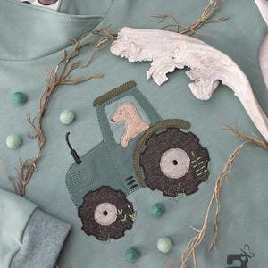 Puede incluir: Una sudadera con capucha verde con un lindo diseño bordado de un tractor con un perro dentro. El tractor está rodeado de hojas verdes y pequeñas bolas de algodón blancas. La sudadera tiene una pequeña máquina de coser bordada con la palabra "Lala" en ella.