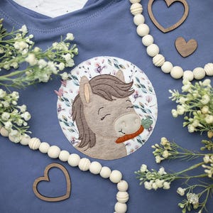 Könnte beinhalten: Blaues T-Shirt mit einem kreisförmigen Applikation eines lächelnden Pferdes mit einer Karotte. Das Pferd ist von einem floralen Muster umgeben. Holzherzformen und eine Perlenkette sind um das T-Shirt angeordnet.
