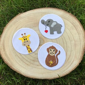 Op de afbeelding: Drie geborduurde strijkpatches met een giraffe, een olifant en een aap. De patches zijn wit met kleurrijke borduursels en liggen op een houten oppervlak.