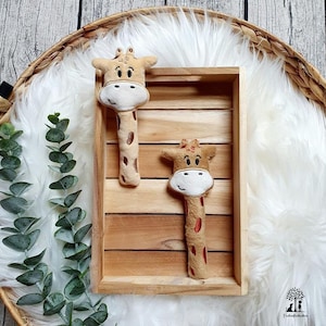 以下が含まれることがあります： 刺繍のディテールが施された、茶色とベージュのキリンのぬいぐるみが2つ。キリンの頭は白で、黒い目と黒い鼻をしています。キリンは白いふわふわの背景のついた木製のトレイの中に置かれています。