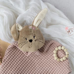 May include: A pink waffle weave baby blanket with a brown bunny plush toy attached. The bunny has a floral headband and embroidered whiskers.