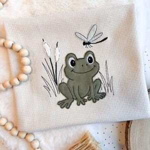 Könnte beinhalten: Ein cremefarbenes, strukturiertes Stoffquadrat mit einer gestickten Fröschin, Schilf und einer Libelle. Der Frosch ist grün mit schwarzen Augen, und die Libelle ist schwarz und weiß. Das Design erinnert an eine skurrile, von der Natur inspirierte Ästhetik.