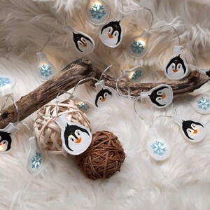 Könnte beinhalten: Eine Lichterkette mit weißen LED-Lichtern mit Pinguin- und Schneeflockenmotiven. Die Lichter sind auf einem weißen Kabel aufgefädelt und leuchten. Die Lichter liegen auf einer weißen, flauschigen Oberfläche.