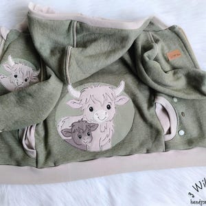 Könnte beinhalten: Ein grüner Kapuzenpullover mit Reißverschluss, hellbeigem Besatz und Kapuze. Der Kapuzenpullover hat aufgestickte Aufnäher einer Hochlandkuh und ihres Kalbes. Der Text "3 Wilde Kerle handgemachte Unikate" ist unten rechts zu sehen.