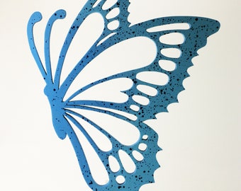May include: A blue butterfly wall decor with a speckled finish. The butterfly is made of wood and has a cutout design. The butterfly is mounted on a clear acrylic stand.