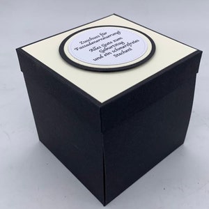 May include: A black and white gift box with a white label on top. The label has a black circle with the text "Zuschuss f&uuml;r Fassadenerneuerung! Alles Gute zum Geburtstag und ein schmerzfreies Stechen!"