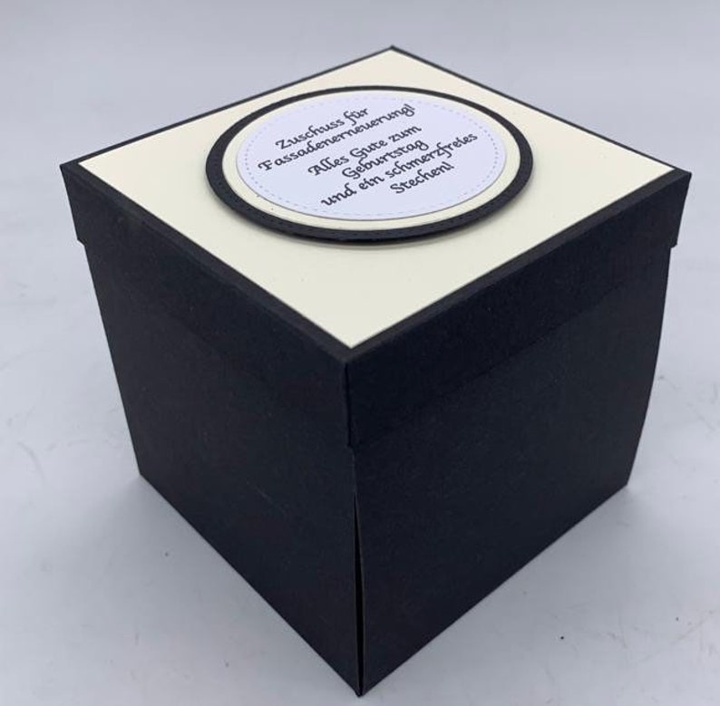 May include: A black and white gift box with a white label on top. The label has a black circle with the text "Zuschuss f&uuml;r Fassadenerneuerung! Alles Gute zum Geburtstag und ein schmerzfreies Stechen!"