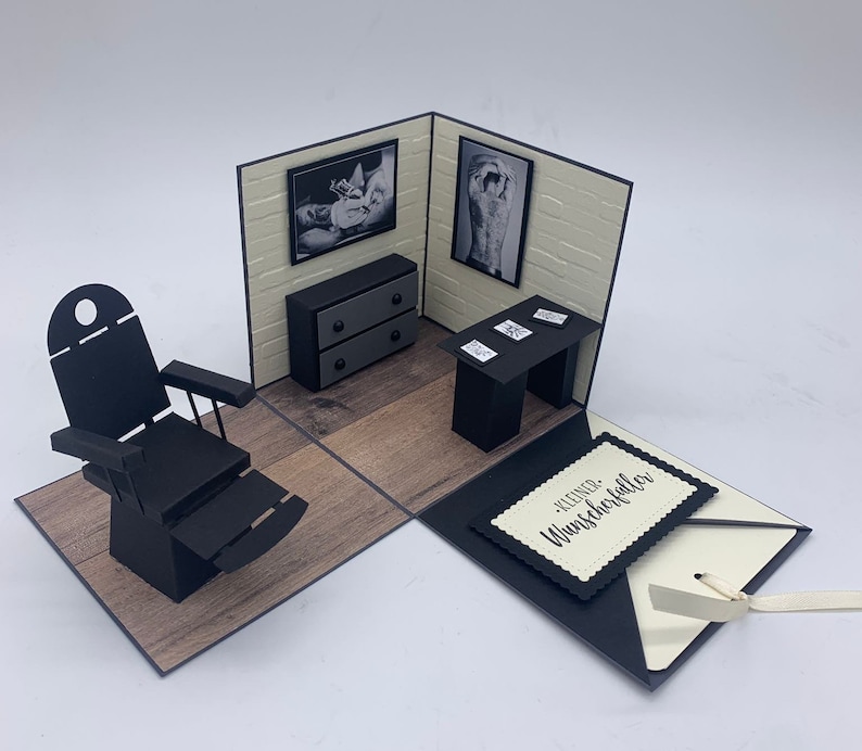 May include: A black and white pop-up card with a tattoo parlor theme. The card features a black leather chair, a small desk with a lamp, and a framed photo of a tattoo artist working on a client. The card is open and the inside reads "Kleiner Wunschzettel f&uuml;r".