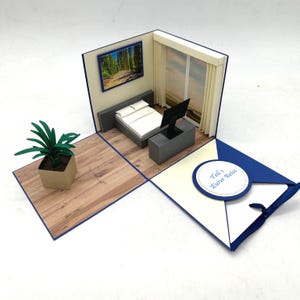 Könnte beinhalten: Handgefertigte, offene Karte mit einer Miniatur-Schlafzimmerszene. Die Szene umfasst ein Bett, einen Fernseher und ein Landschaftsgemälde. Eine kleine Topfpflanze steht auf einem Holzboden. Die Karte ist weiß und blau, mit dem Text "Teil 1 Eurer Reise".