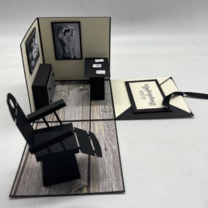 May include: A black and white 3D pop-up card with a barber chair, a table with a black top, and a picture of a man with a tattoo on his back. The card is open and the inside is decorated with a wood-grain pattern. The card has a black tag with the text "Alles Gute zum Geburtstag" on it.