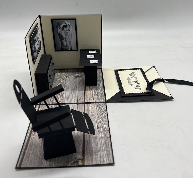 May include: A black and white 3D pop-up card with a barber chair, a table with a black top, and a picture of a man with a tattoo on his back. The card is open and the inside is decorated with a wood-grain pattern. The card has a black tag with the text "Alles Gute zum Geburtstag" on it.