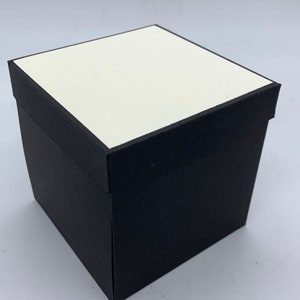 May include: A black square box with a white top. The box is made of cardboard and has a smooth finish.