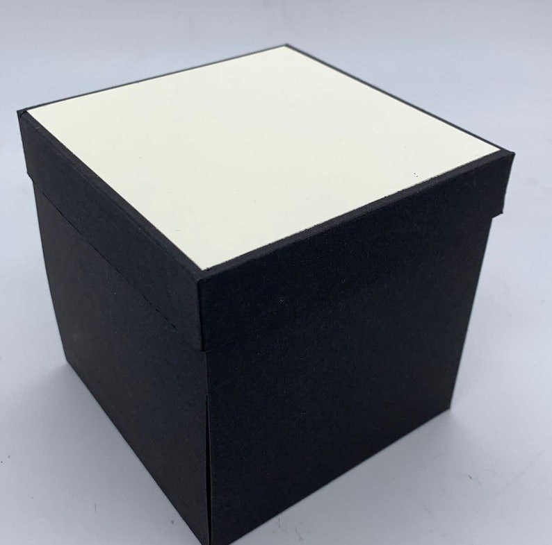 May include: A black square box with a white top. The box is made of cardboard and has a smooth finish.