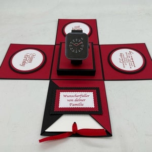 Könnte beinhalten: Eine rote und schwarze Explosionsbox mit einer schwarzen Uhr im Inneren. Die Box hat eine weiße Karte mit dem Text "Wunscherfueller von deiner Familie" auf der Vorderseite. Die Box ist mit einem roten Band gebunden.