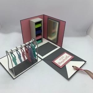Könnte beinhalten: Eine rosa und graue Pop-up-Karte mit einem Miniatur-Kleiderladen im Inneren. Der Laden hat einen Kleiderständer mit bunten Kleidern, ein Regal mit gefalteten Kleidern und einen Spiegel. Die Karte hat ein rosa Etikett mit dem Text "kleiner Wunsch erfüllt".