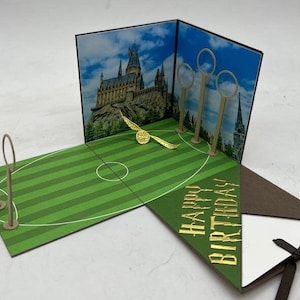 Könnte beinhalten: Eine Pop-up-Karte mit einem grünen Feld, drei goldenen Reifen und einem goldenen Schnatz, der durch die Luft fliegt. Der Hintergrund zeigt Hogwarts-Schloss. Die Karte ist geöffnet und innen steht "Happy Birthday".