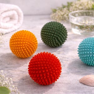 Könnte beinhalten: Vier kugelförmige Massagebälle mit einer texturierten, stacheligen Oberfläche. Das Set enthält einen orangefarbenen, einen dunkelgrünen, einen leuchtend roten und einen hellblauen Ball. Jeder Ball hat ein gleichmäßiges Muster aus kleinen Spitzen.